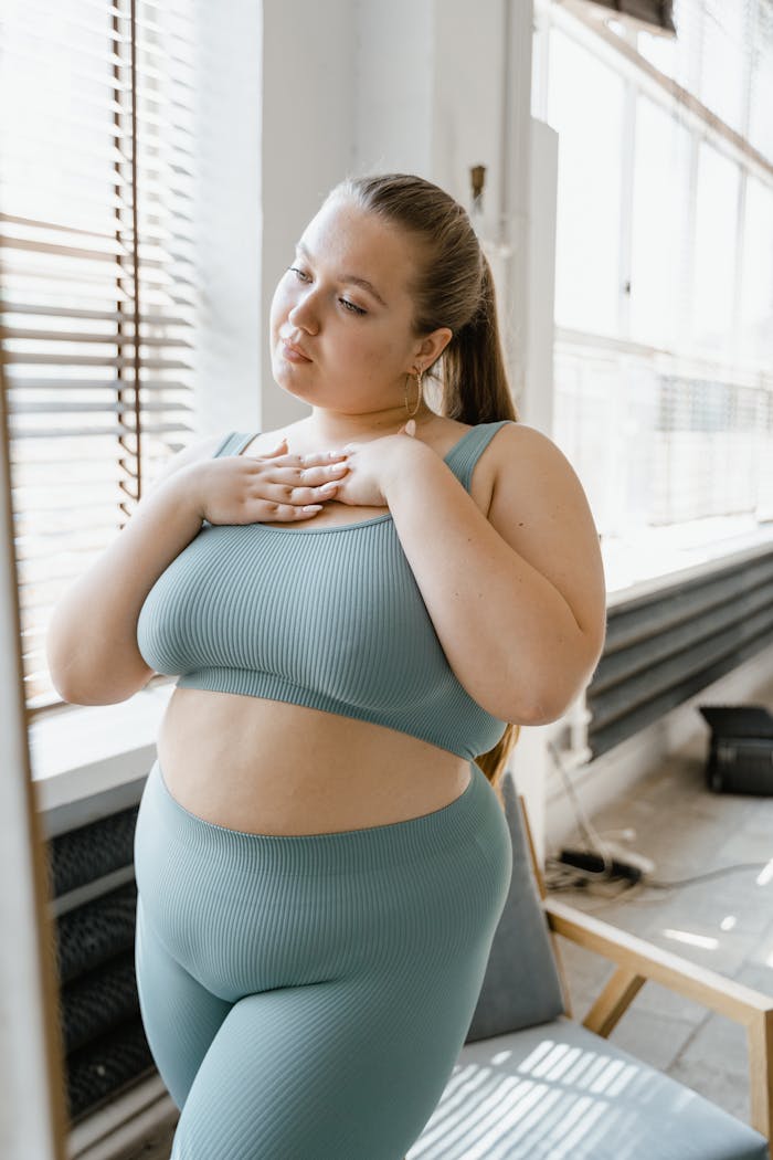 Plus-size woman in teal leisurewear standing by a window, exuding confidence and self-esteem.
